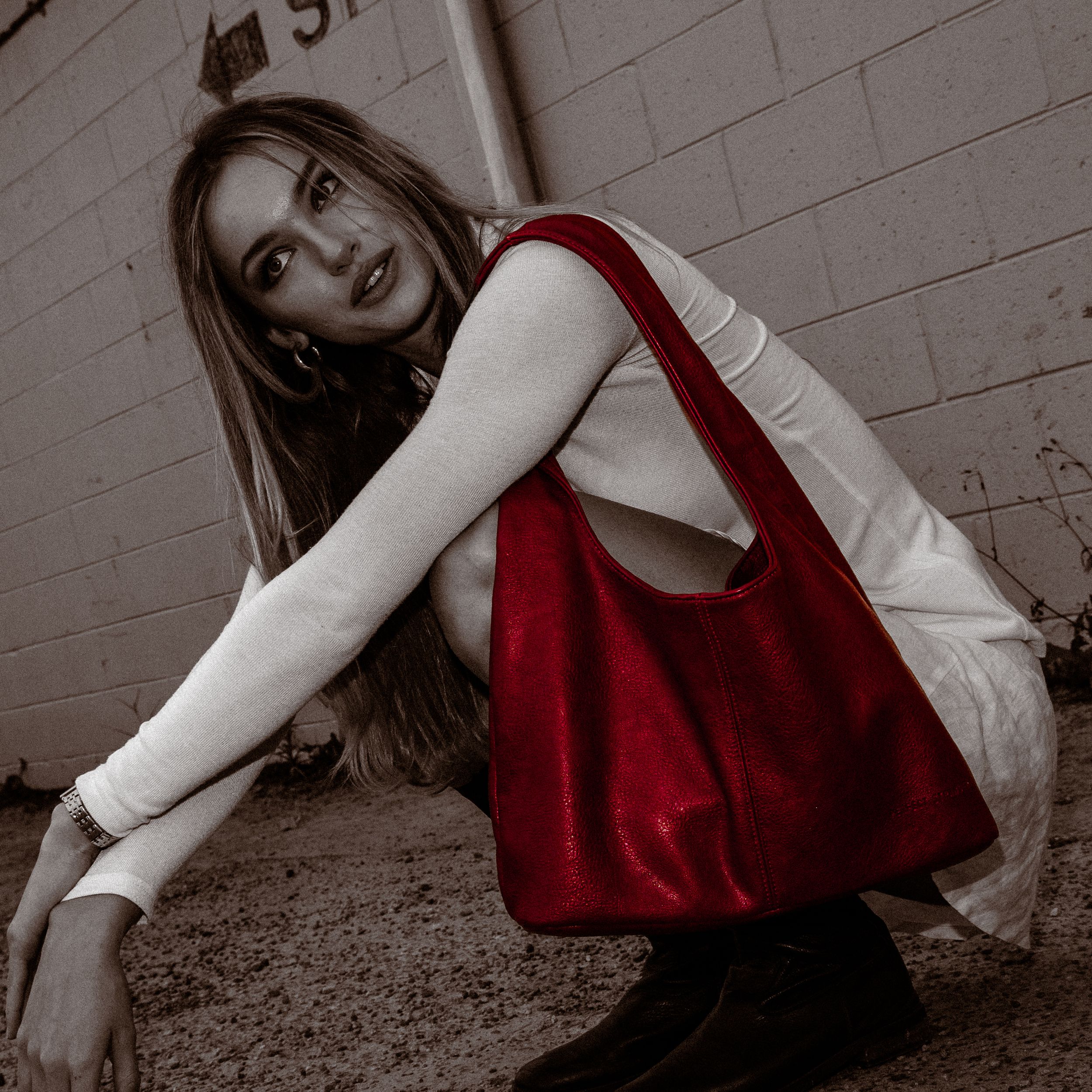 A person in a red dress and white sweater crouches against a brick wall in a moody monochromatic composition.