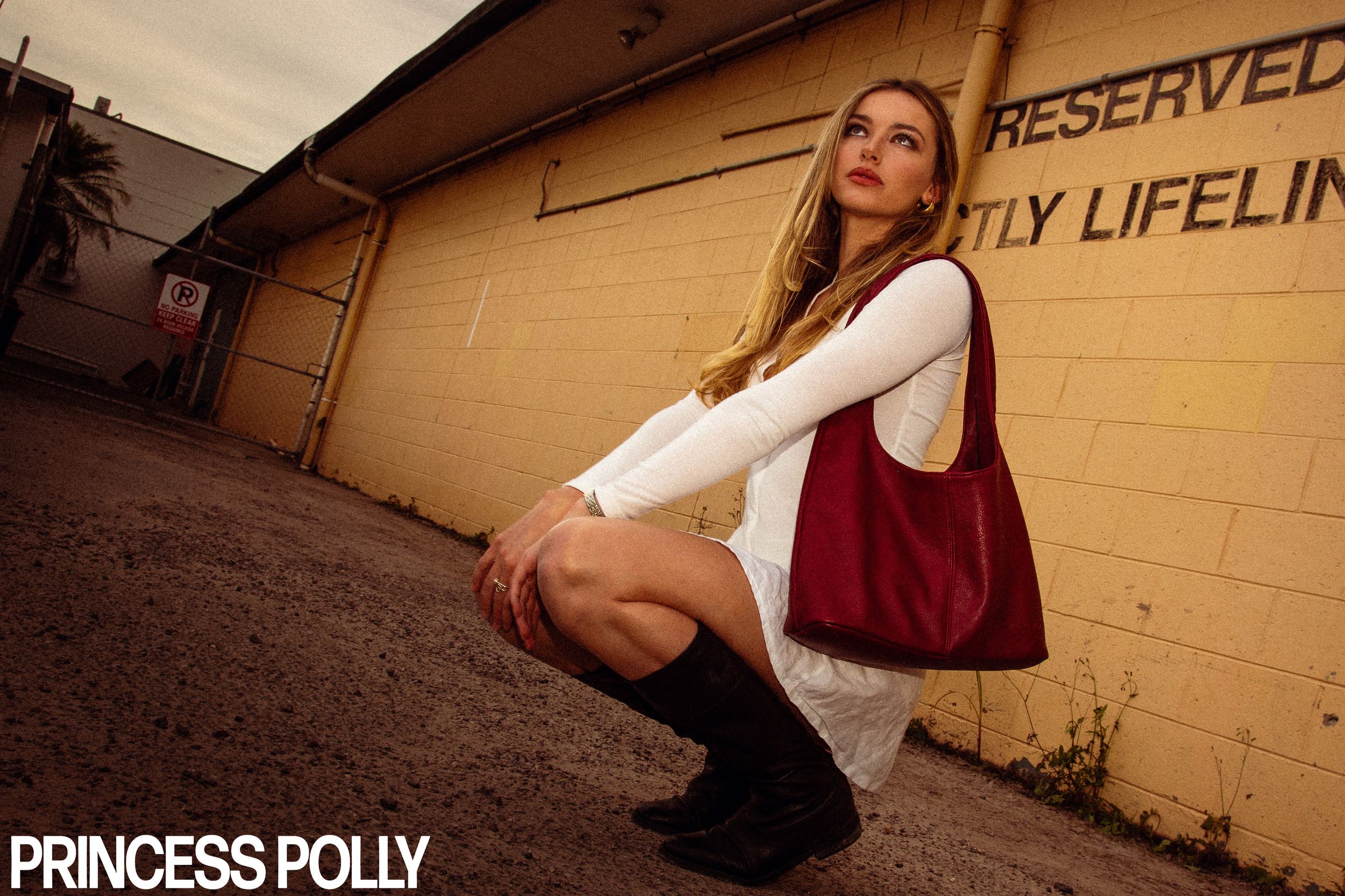 Low angle photo of someone in white dress and burgundy vest crouching against a beige wall with Reserved text.