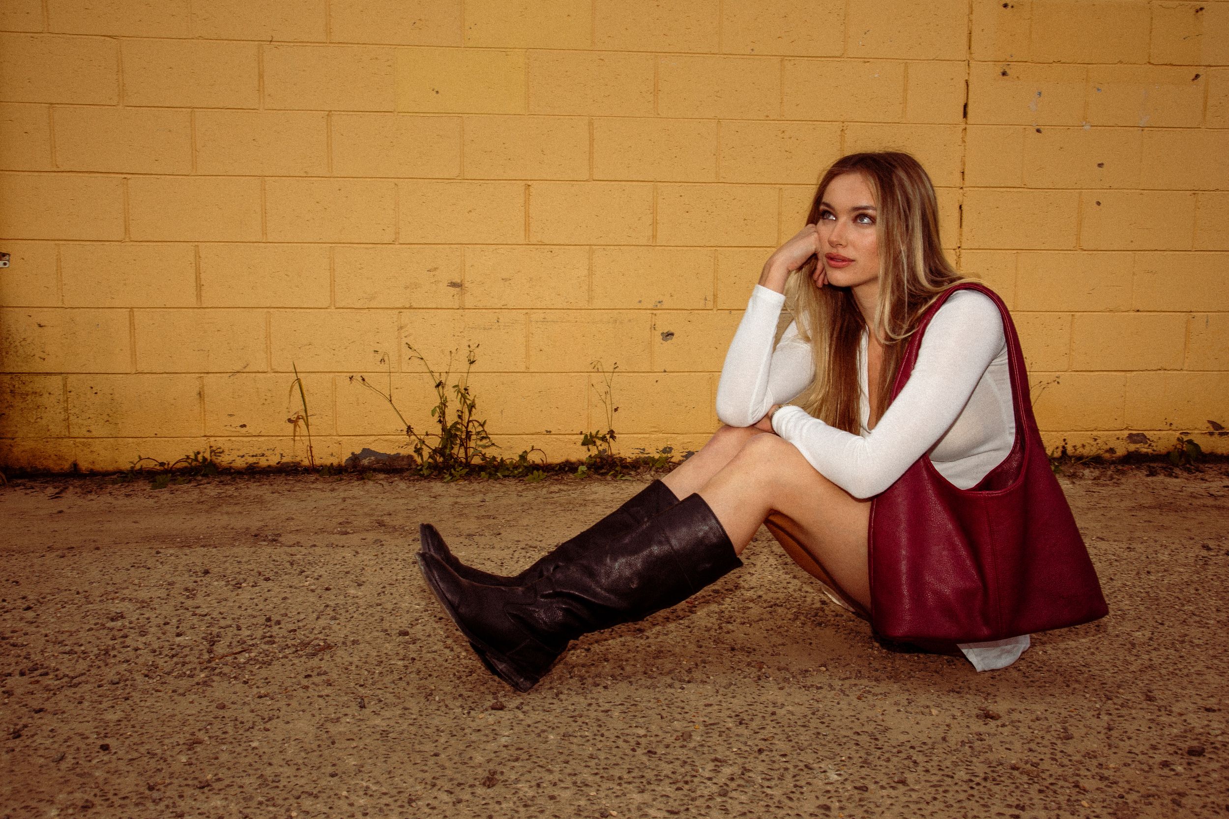 Figure in a burgundy dress sits thoughtfully on a white brick wall as golden sunlight illuminates the scene.