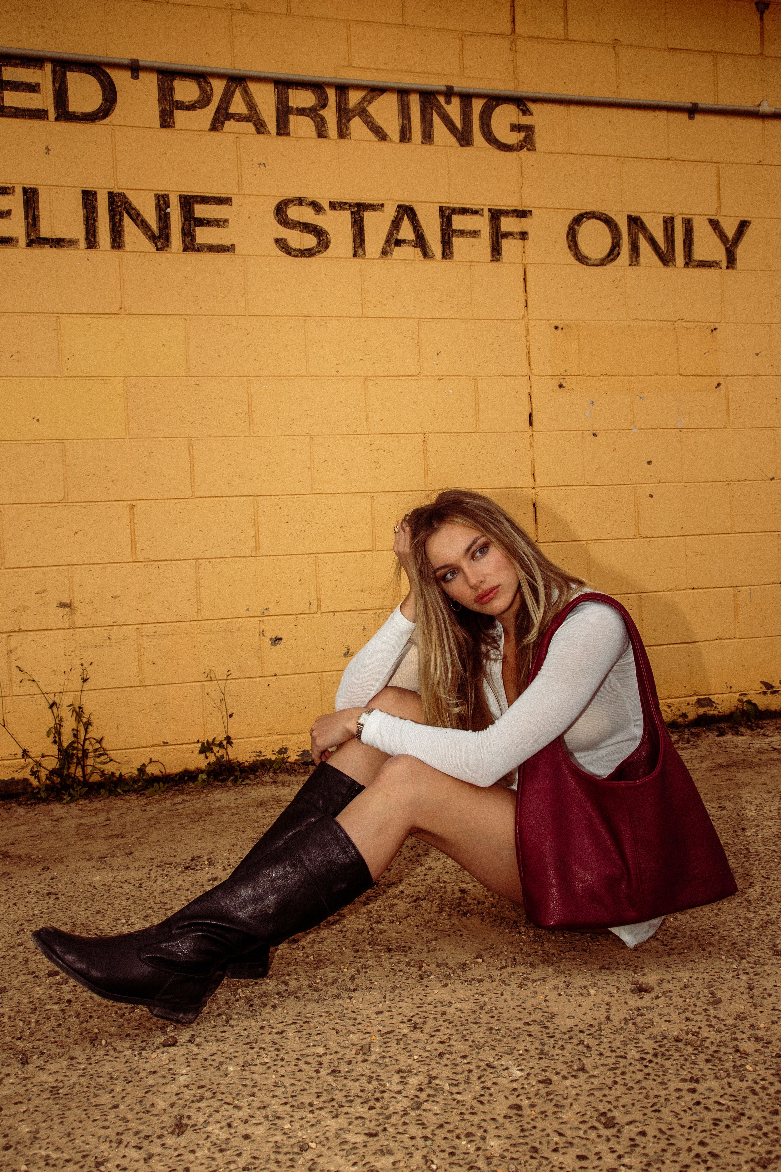 Someone in a burgundy dress with boots sits against a yellow brick wall near a reserved parking area at dusk.