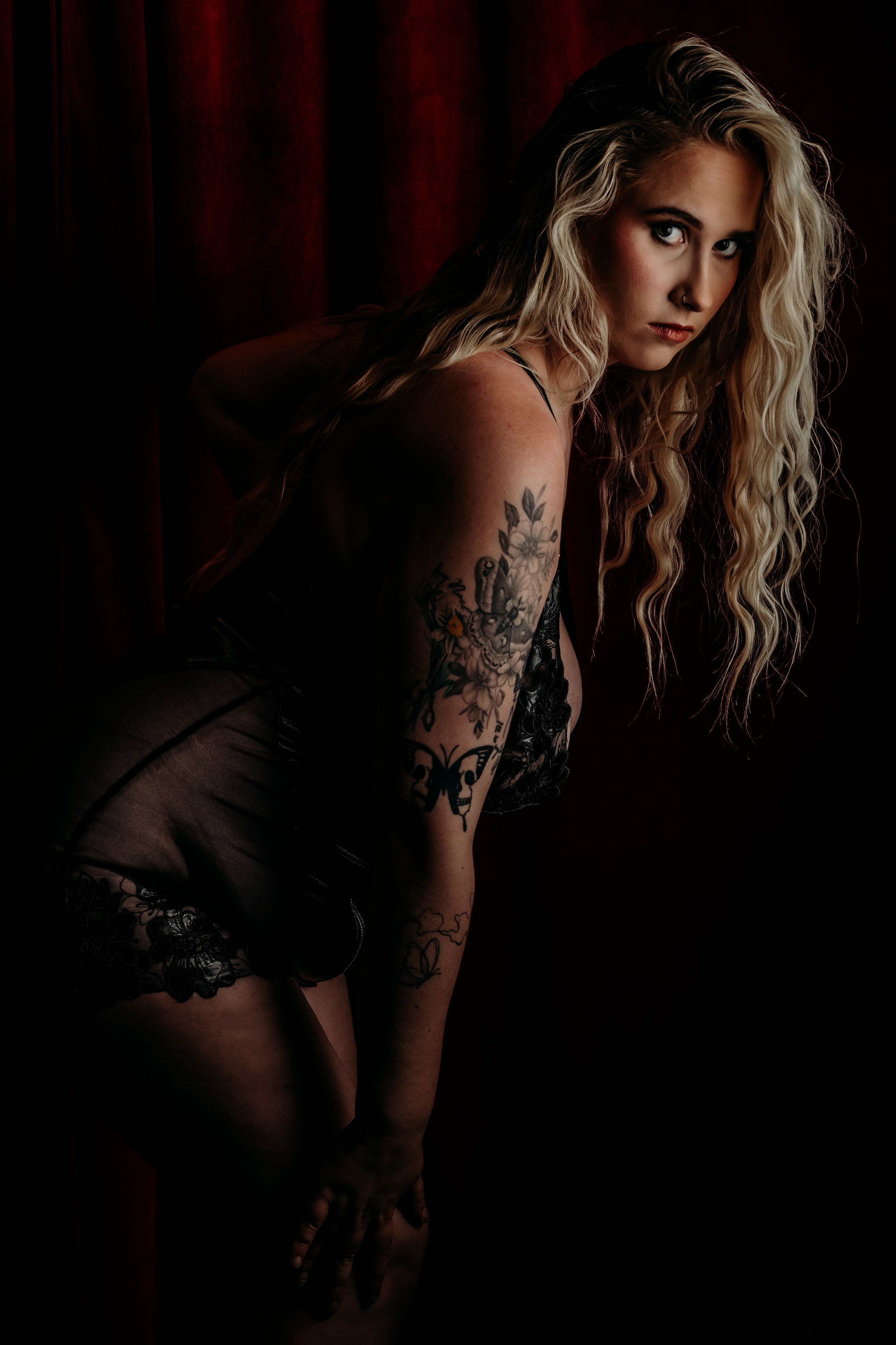Moody boudoir portrait with dramatic red backdrop showing blonde model in black lingerie with floral arm tattoos. grand rapids boudoir MI