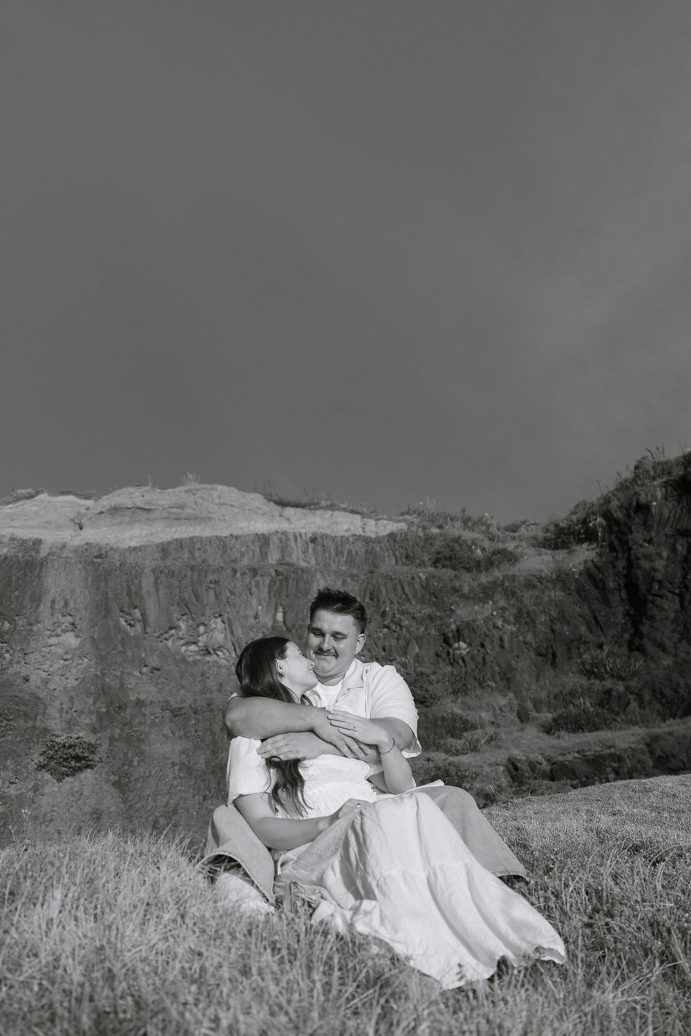 A couple lying together on a grassy hillside overlooking the ocean in black and white.