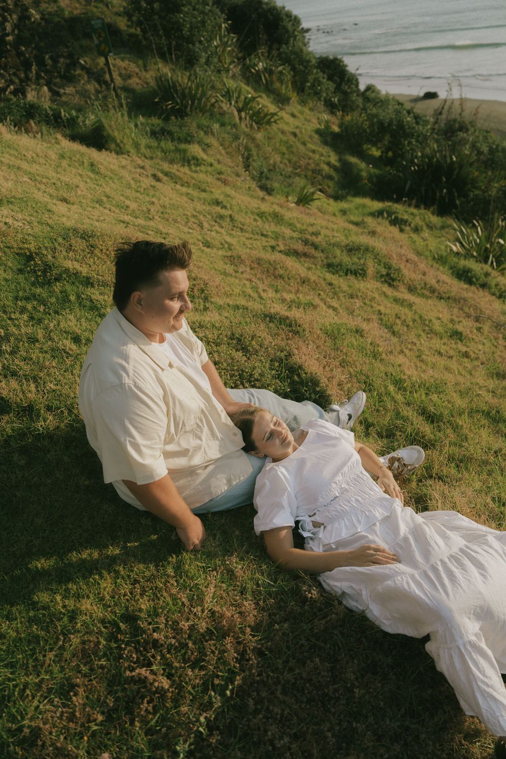 A couple resting on a grassy hillside in white clothing during golden hour.