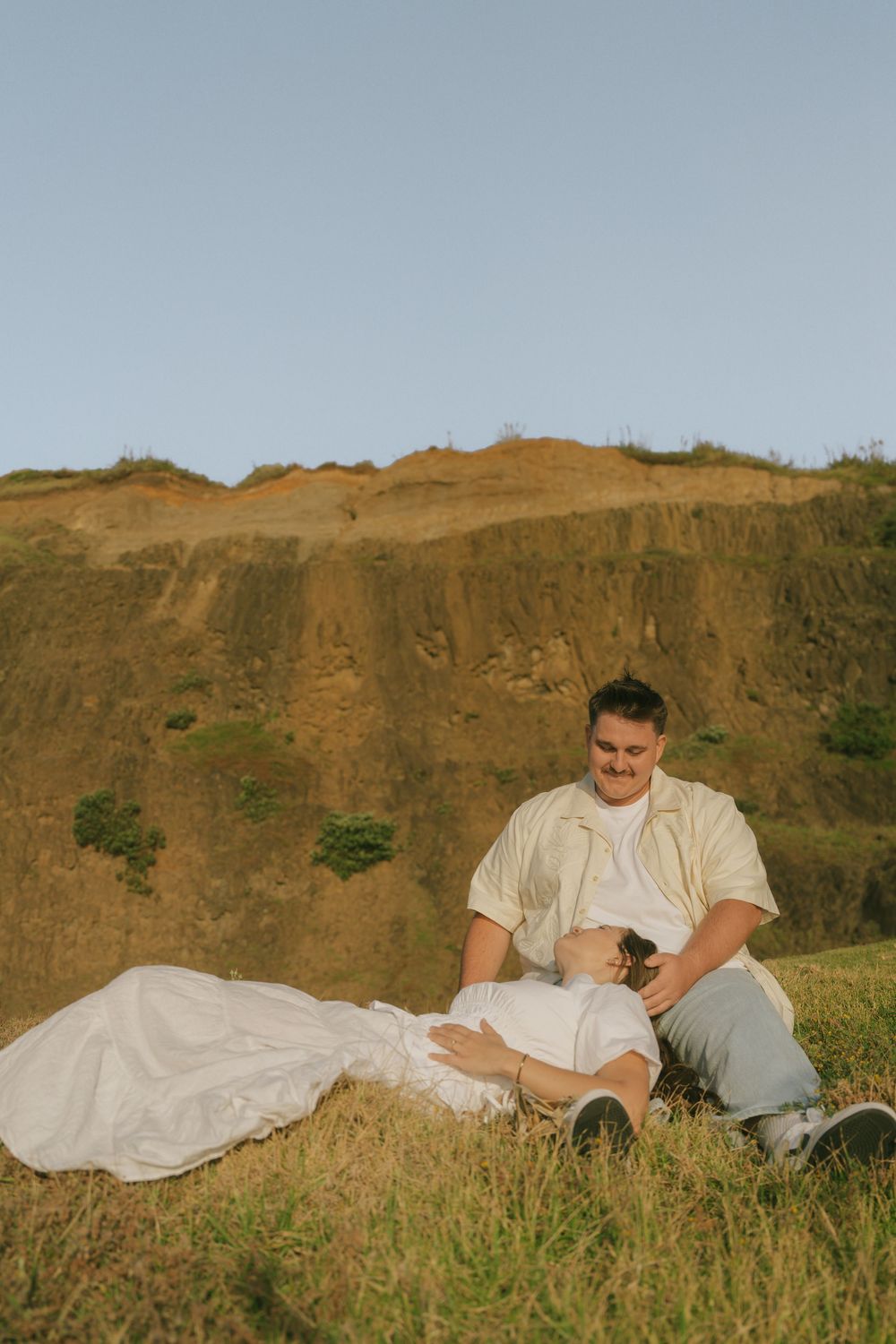 A couple relaxing on a grassy slope against a backdrop of coastal cliffs.