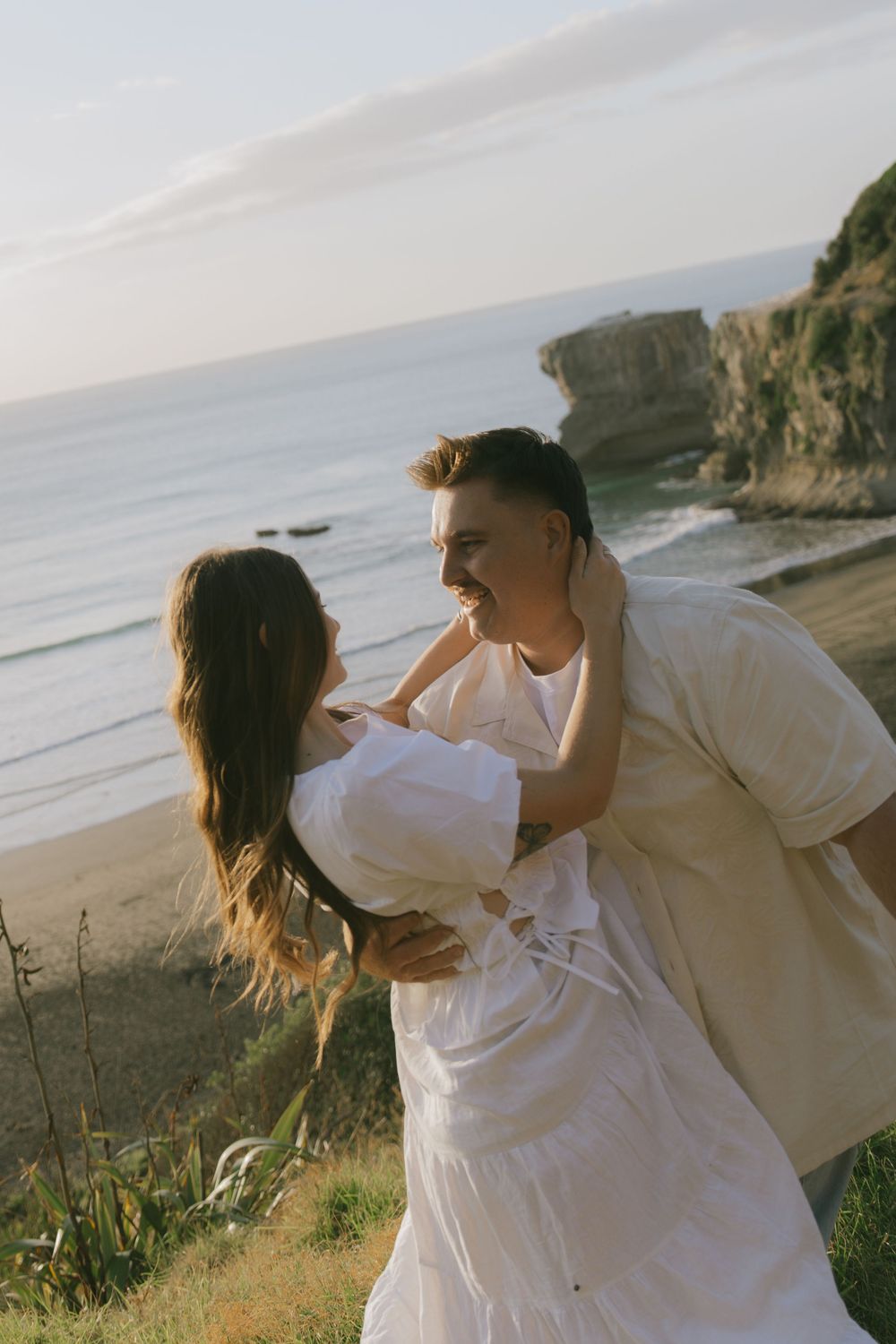 A playful couple in white clothing embracing on a coastal cliff at sunset.