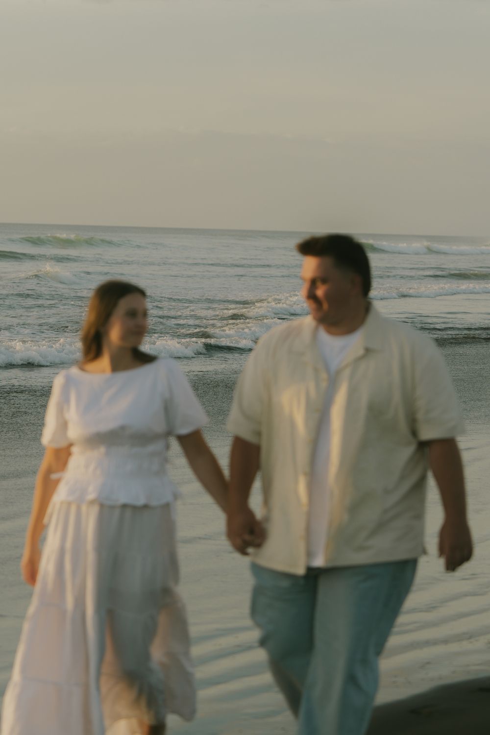 A couple in white clothing walking hand in hand along a beach at sunset.