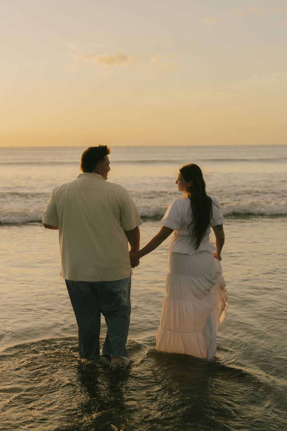 Two people in white clothing wade together in ocean water during sunset.