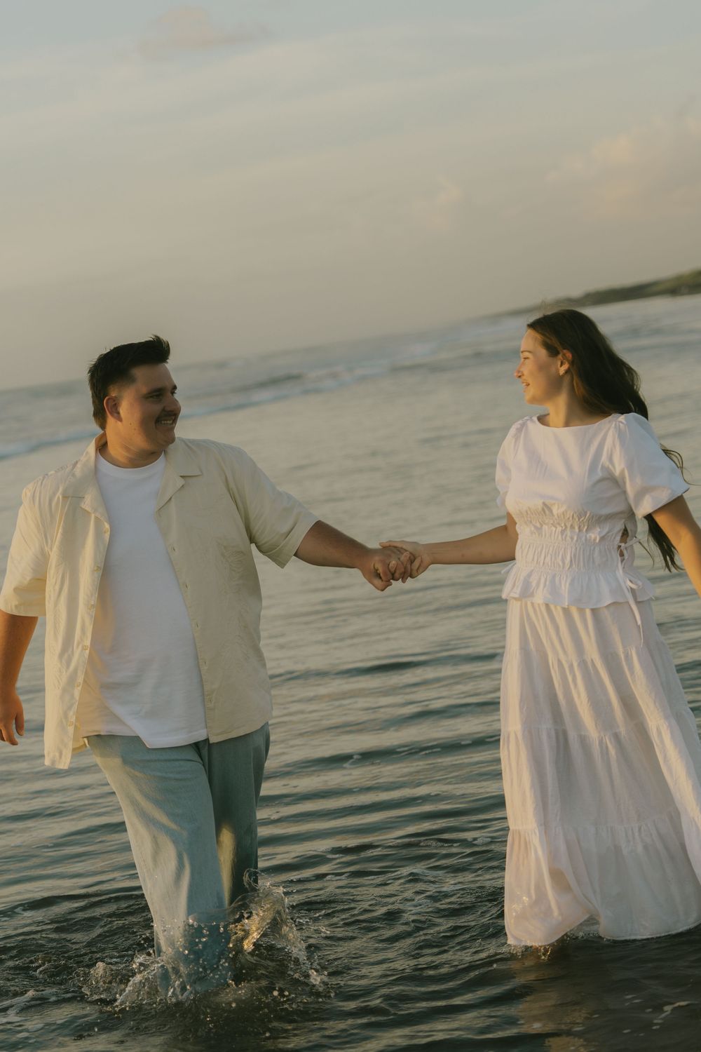 A couple holding hands while wading through ocean waves at sunset.