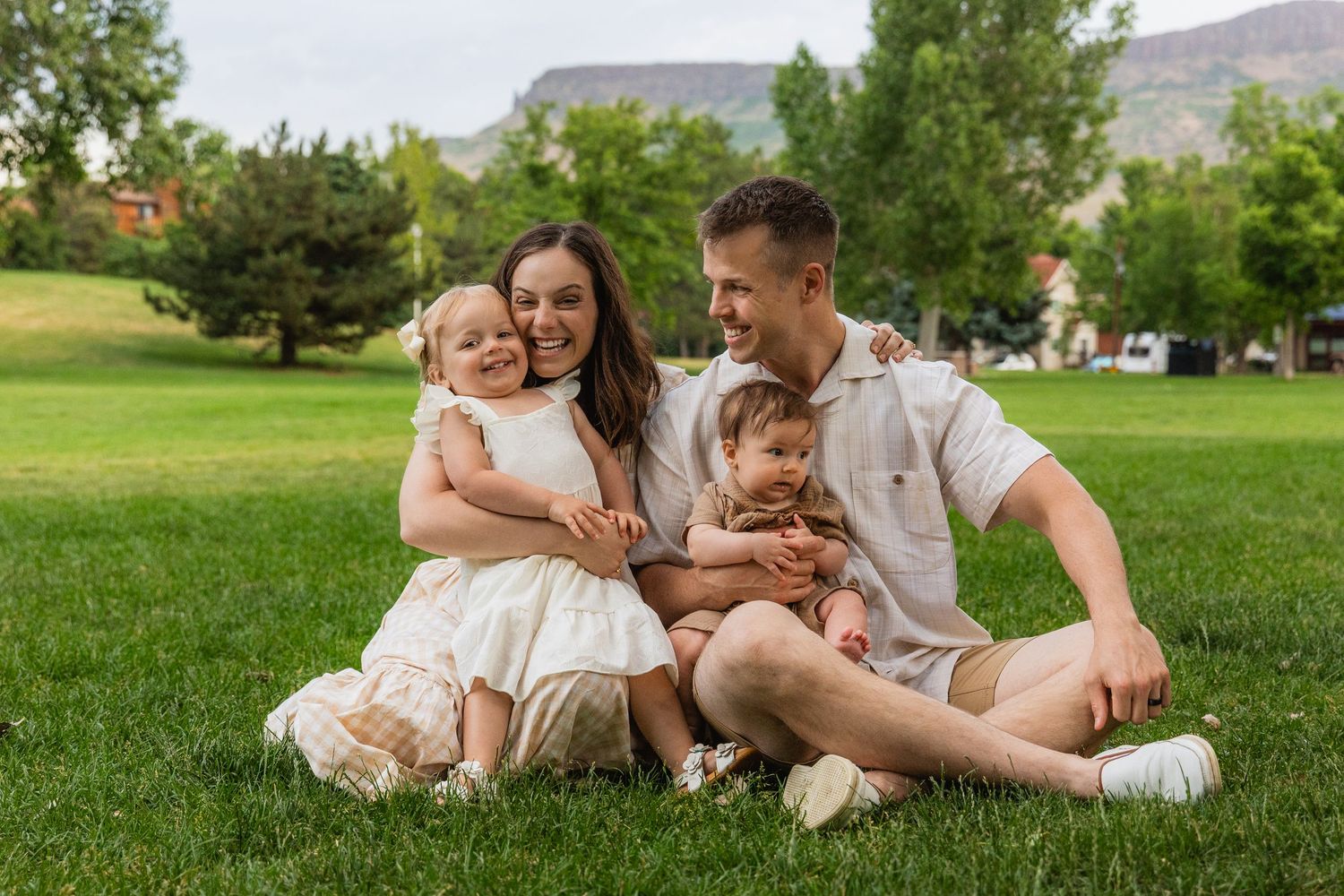 A family sits together on lush green grass with mountains and homes visible in the background on a sunny day.