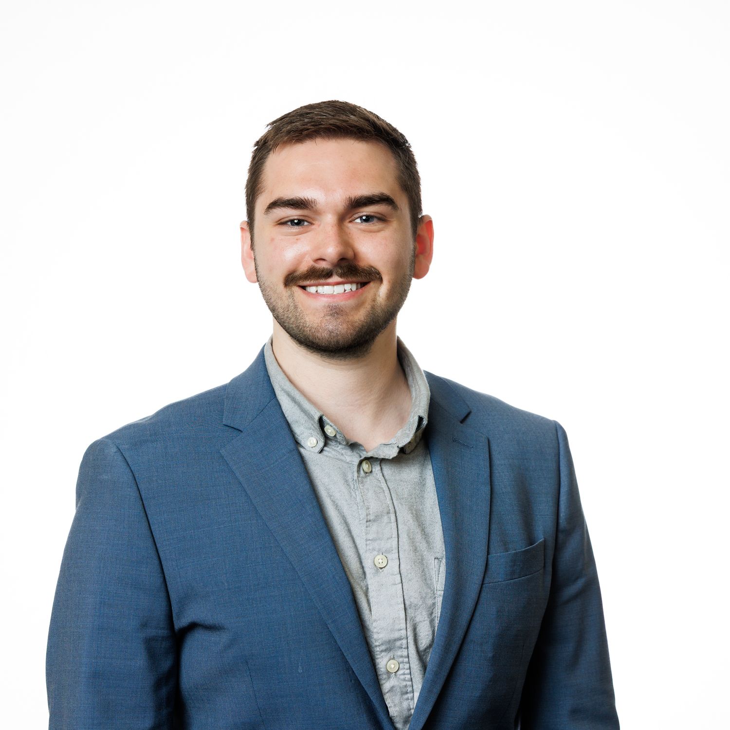Professional headshot of someone in a blue blazer and grey button-up shirt smiling against a white background.