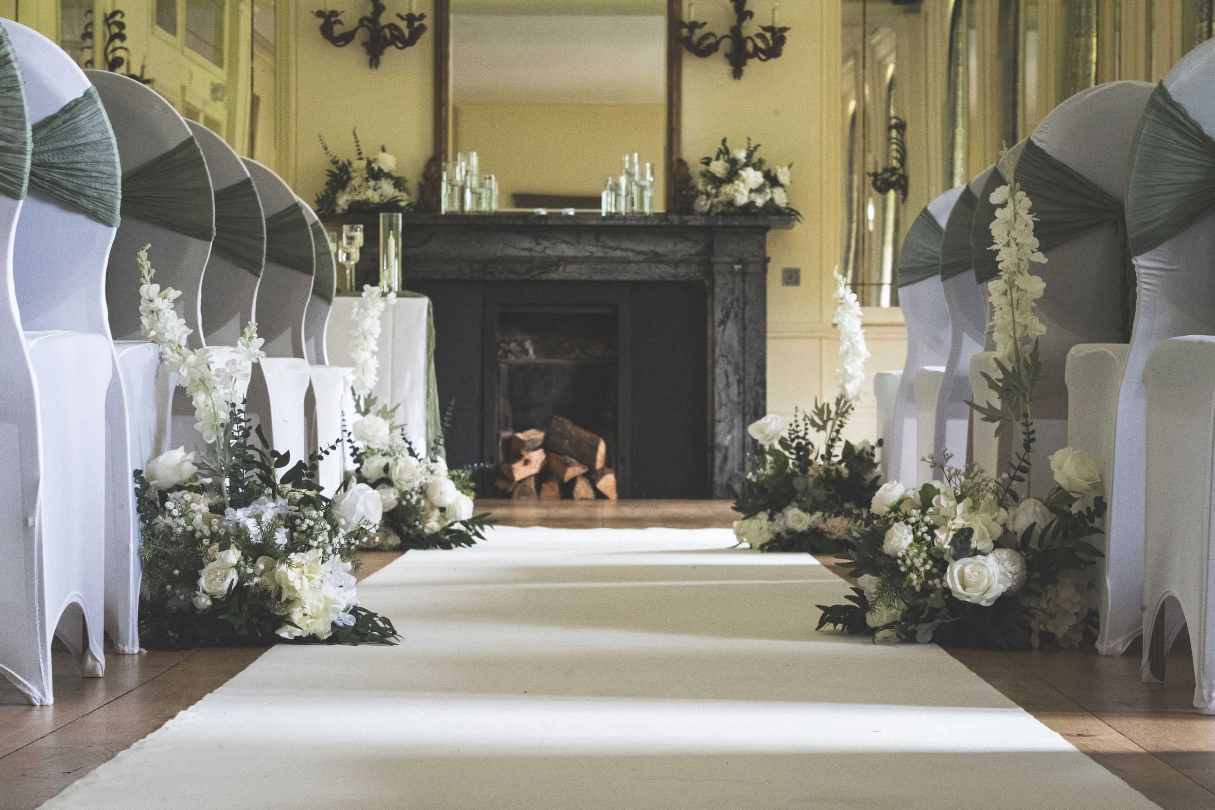 White wedding ceremony aisle with chair covers and floral arrangements leading to an elegant fireplace in a historic venue.
