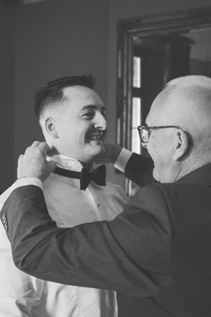 A black and white photograph shows someone adjusting another person's bowtie in an intimate pre-wedding moment.