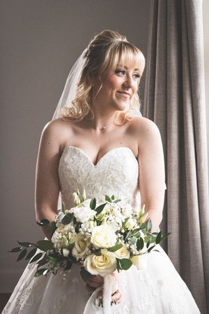 A bride in a white strapless lace gown holds an elegant cream and white rose bouquet while standing near window drapes.