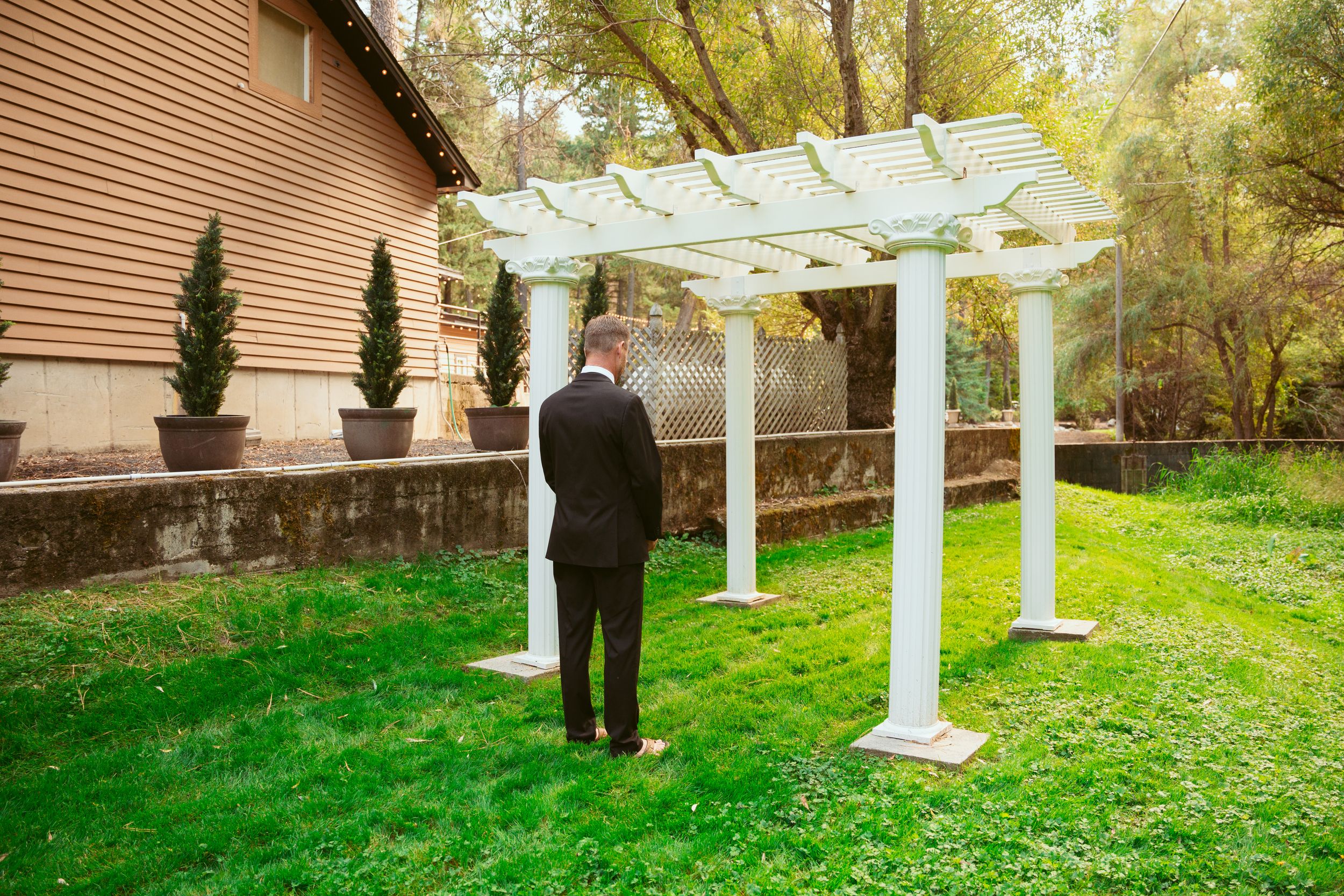 A picturesque outdoor pergola stands against a beige house with lush green grass and autumn foliage in the background.