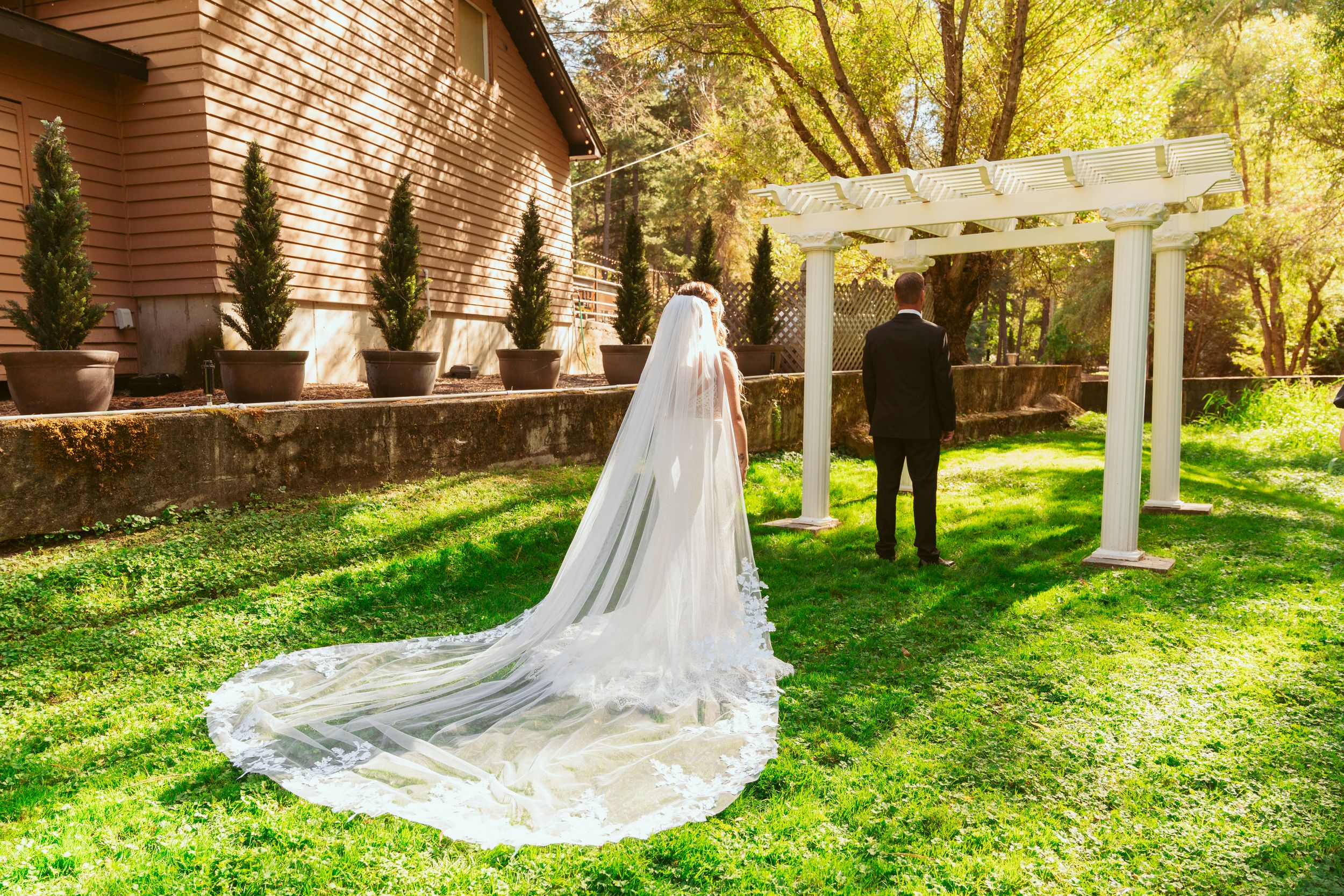 A long flowing wedding veil trails on green grass near a white garden arbor on a sunny afternoon.