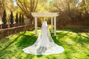 Long flowing wedding dress and veil photographed from behind under a white garden pergola in golden sunlight.