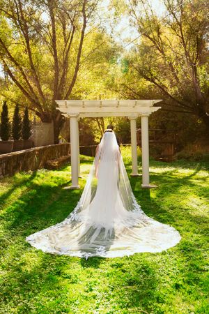 A dramatic wedding gown with flowing cathedral veil displayed under a white garden pergola in golden sunlight.
