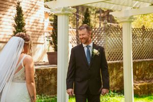 Beautiful outdoor garden wedding ceremony features a bride in white veil and someone in dark suit under a white arbor.