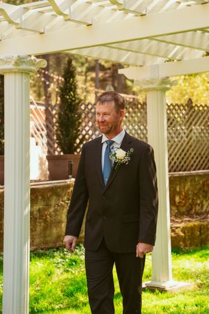 Figure in a black suit and blue tie stands under a white garden pergola during an outdoor wedding celebration.