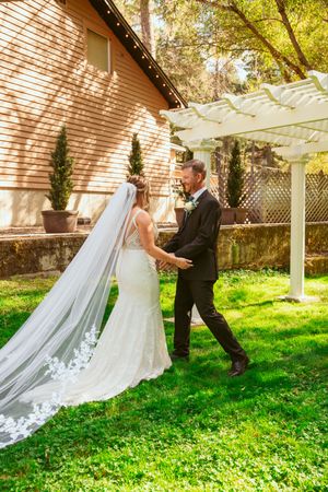 An elegant wedding couple shares a moment together under a white pergola in a sunny garden setting.