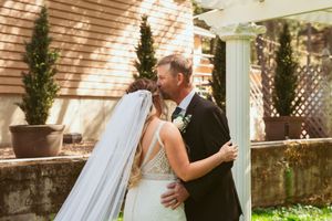 A tender moment captured between newlyweds embracing on their wedding day in a garden courtyard setting with potted cypress trees.