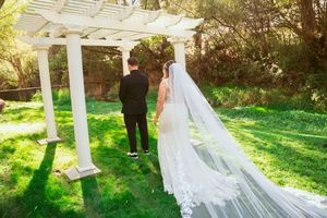 A romantic wedding couple poses together in a sunlit garden setting beneath a white pergola with a flowing veil.
