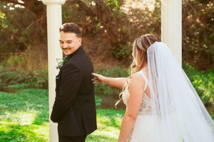 A romantic wedding first-look moment captured in a garden with sunlight streaming through the trees and long white veil.
