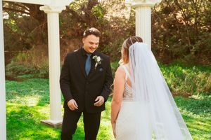 A couple in formal wedding attire stands together outdoors under a sunny sky among green trees.