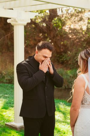 A couple during a first look wedding moment under a white pergola with lush greenery in the background.