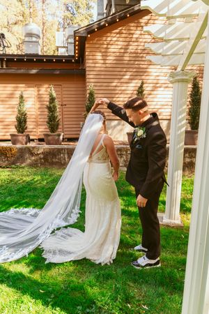 A romantic wedding moment captured under a white pergola beside a rustic wooden building.