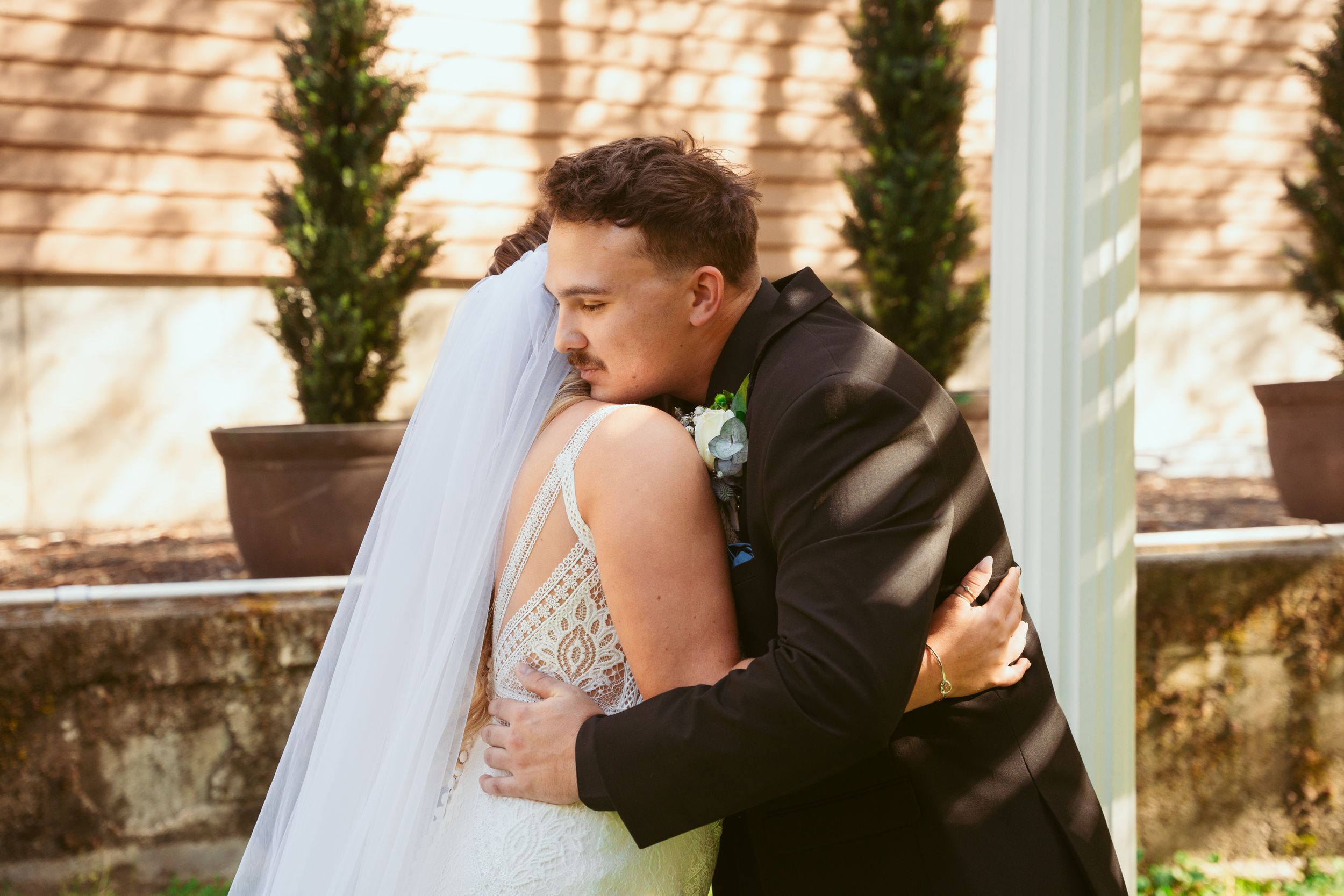 Two people share a tender embrace and kiss in an elegant outdoor venue with white columns and greenery.