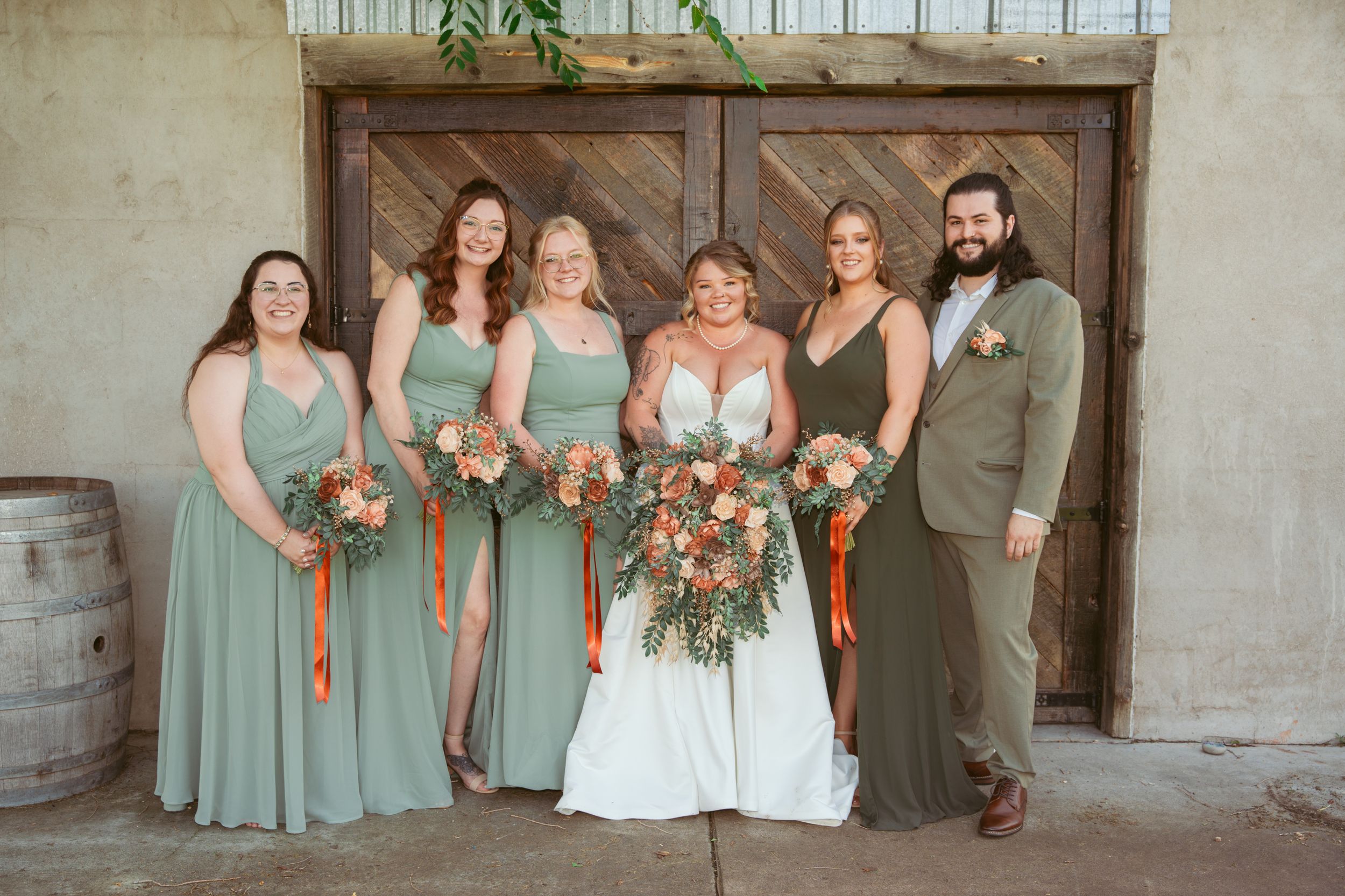 Wedding party in coordinated sage green dresses and formal attire pose on stone staircase for group photo.