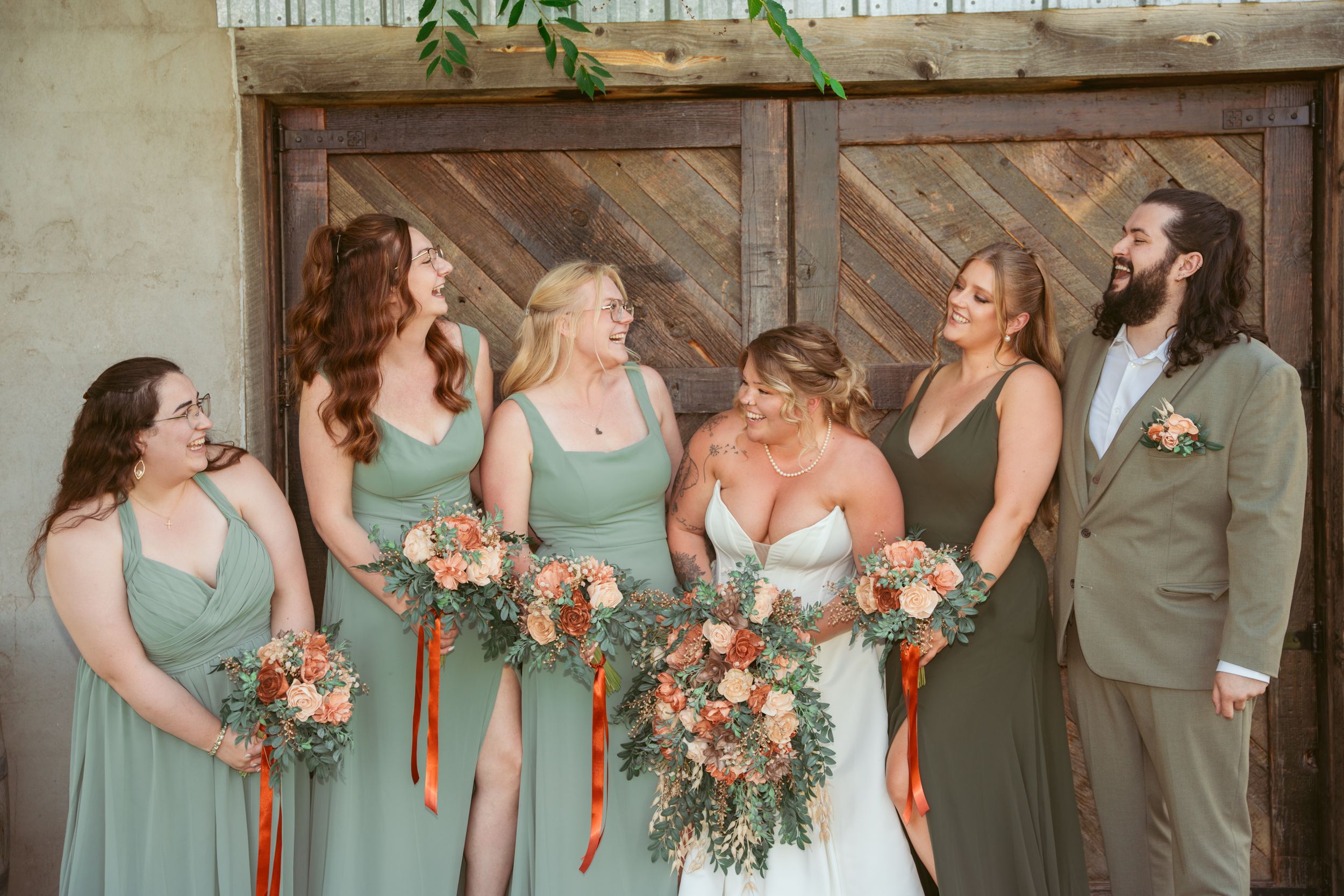 Wedding party dressed in sage green and grey poses and laughs together in front of rustic wooden doors.