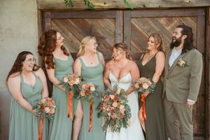Wedding party in sage green dresses and gray suit share a candid laughing moment against rustic wooden barn door.