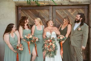 Wedding party dressed in sage green poses together outdoors while holding peach and coral floral bouquets.