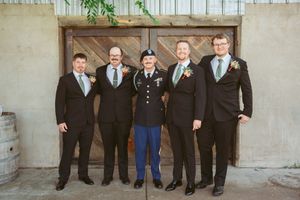Group in formal attire stands together against concrete wall for military wedding photo.