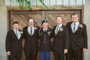 A group of formally dressed wedding guests in dark suits standing together for a portrait with rustic wooden door backdrop.