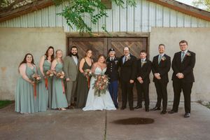 Wedding party members pose together in formal attire on a sunny day outside a rustic white building.