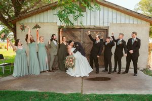 Wedding party celebrates with joy outside rustic barn entrance with bridesmaids in sage green dresses.