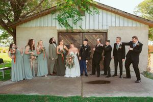 Playful wedding party poses outside white barn with bridesmaids in sage gowns and groomsmen in black suits.