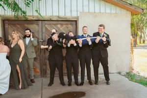 A wedding party poses in line against a rustic beige wall in an outdoor setting with greenery.
