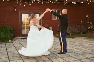 A bride in a flowing white dress dances with her military groom on a patio with string lights overhead.