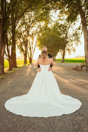 Flowing white wedding dress train spreads out beautifully against a grassy path with sunlit trees in the background.
