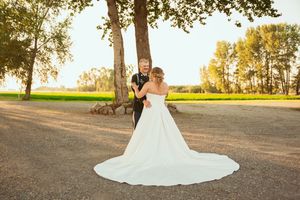 White wedding dress train flows across dirt path with trees and golden sunlight in background.