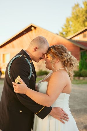 Romantic wedding couple embracing against rustic red barn backdrop in evening light.