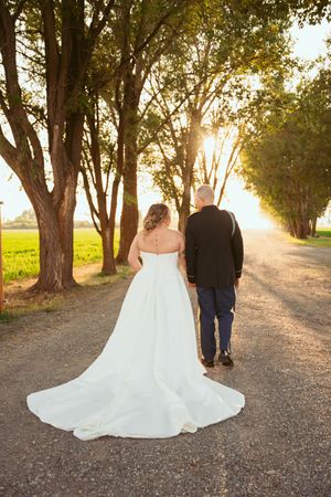 A couple walks down a sun-dappled dirt road lined with trees in wedding attire.
