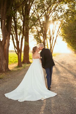 A couple walks down a tree-lined path at sunset in wedding attire.