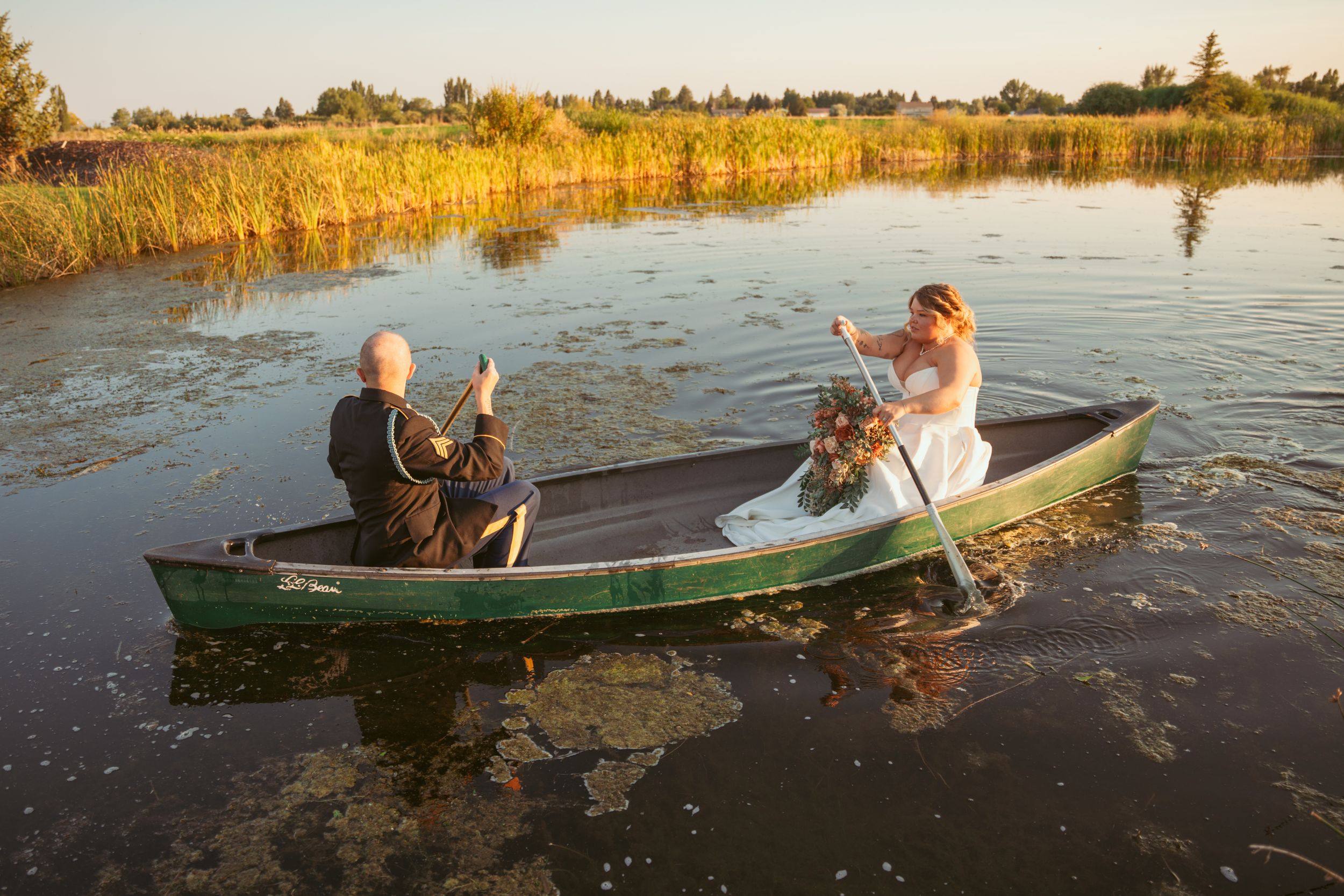 Wedding couple sits in a canoe on a golden-lit lake during sunset with reeds in background.