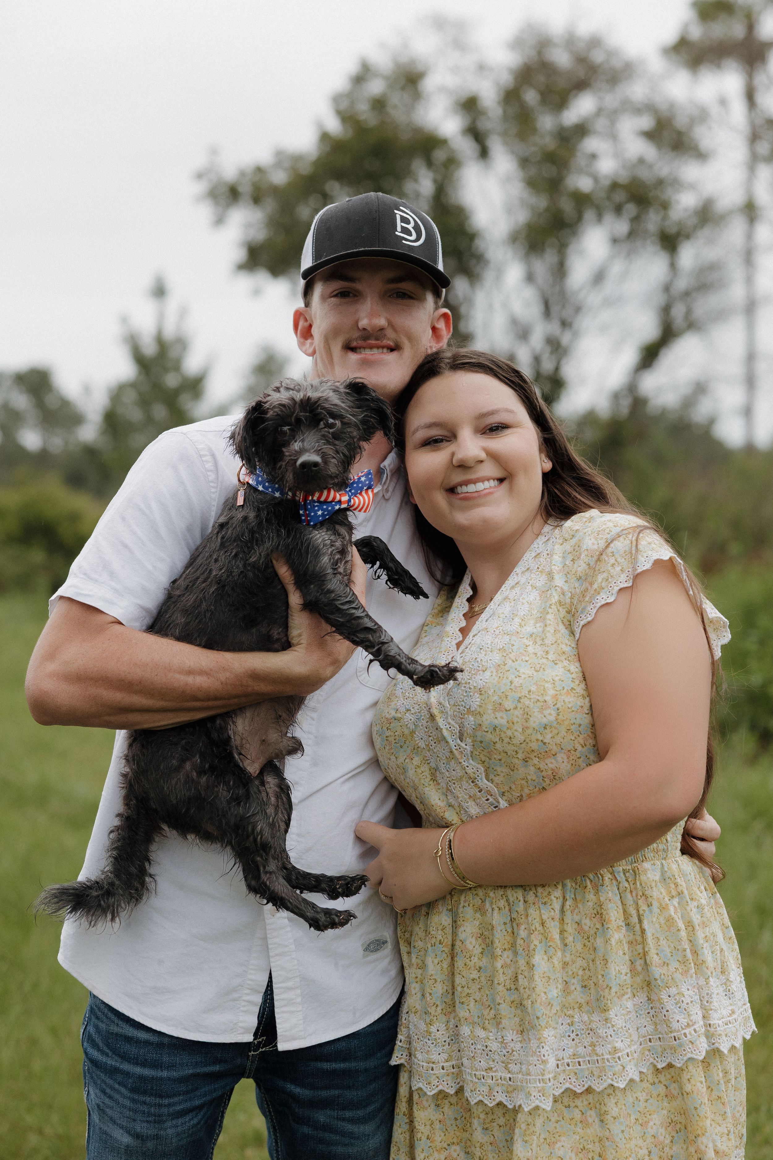 A couple poses with their small black dog in a grassy outdoor setting.
