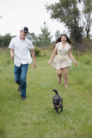 A couple runs and plays with their small black dog in a grassy field.